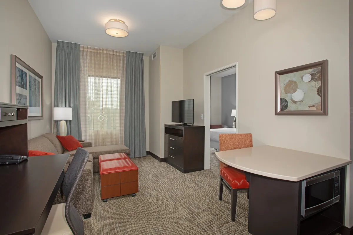 The image showcases a bright, airy living area with a plush sofa adorned with decorative pillows, a sleek dining table set for two, a modern television unit, and a glimpse into the cozy bedroom. Sunlight filters in, creating a warm and inviting atmosphere that feels both relaxing and functional—perfect for unwinding, working, or sharing a quiet meal.