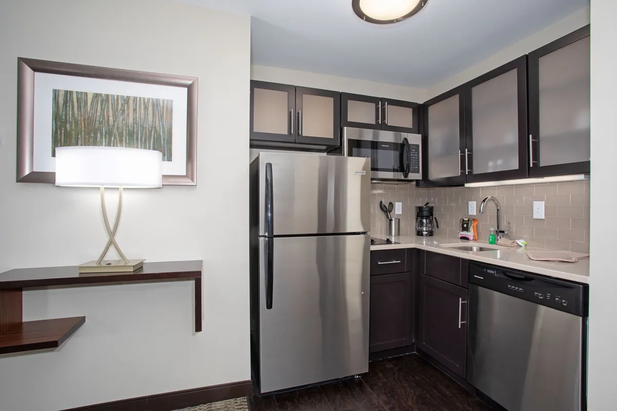 A view of a modern kitchen interior featuring stainless steel appliances, ample kitchen cabinets, a table lamp, and wall art. The inviting space is designed for both practical cooking and social gatherings, with visible utensils and surfaces ready for meal prep or casual dining.