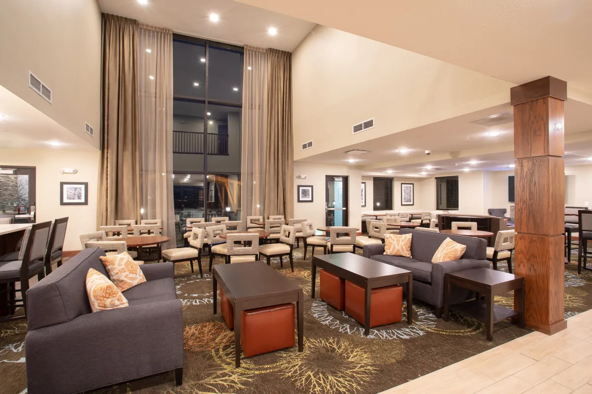 This image depicts a bright, inviting interior lounge area with large windows draped with curtains, multiple seating arrangements, wooden pillars, and artwork adorning the walls. It offers a spacious, comfortable environment that can effortlessly serve both relaxation and informal work needs.