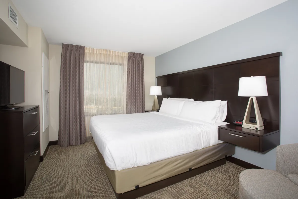 A spacious bedroom featuring a king-sized bed dressed in crisp linens, a large window that floods the room with natural light, a nightstand with a lamp for relaxing evenings, a comfortable accent chair, and a television for unwinding. The space is styled with subtle decorative touches, creating a serene and inviting atmosphere.